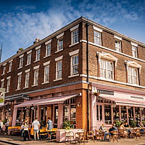 A corner shot of a Georgian Town House style building with sash windows. The ground level of the building has been turned into a cafe, painted a complimentary light pink with awnings and outdoor seating. The sky is blue and it looks like a warm summer day.