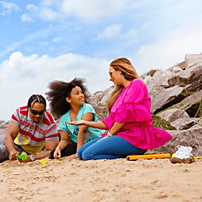 A family are on a white sandy beach in Wirral. The sky is a light blue with some clouds. They are building sandcastles next so some rocks.
