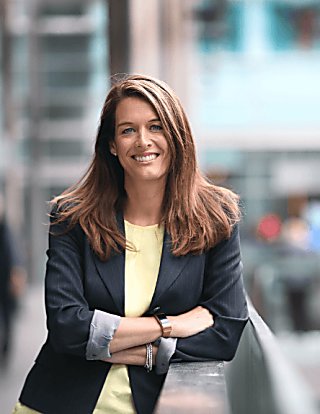 A woman smiling brightly wearing a black blazer with a pale yellow dress, she is looking directly at the camera and smiling while leaning against a wall.