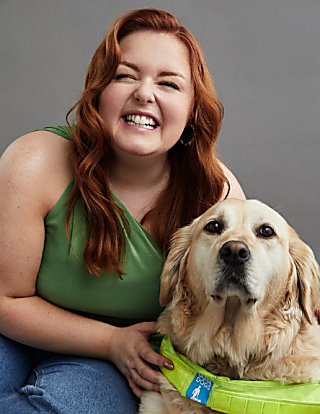 A lady with red, waved hair smiles at the camera whilst holding onto a sandy coloured, golden retriever who is her guide dog.