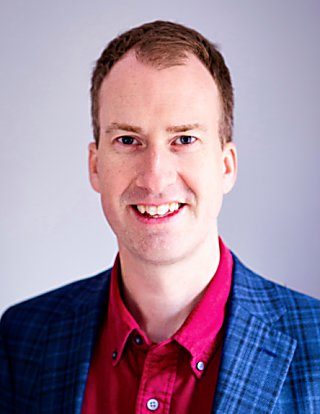 A man wearing a red shirt and blue, tartan suit jacket smiles at the camera.
