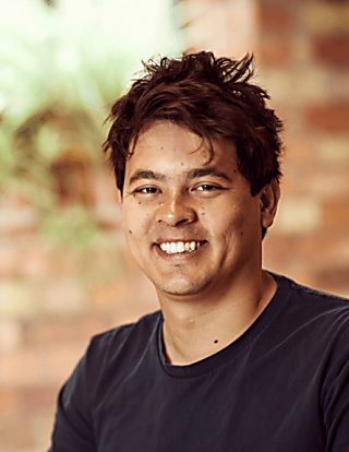 An image of a man smiling brightly and showing his teeth, his hair is black and he is wearing a navy top.