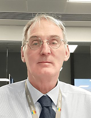 A man looking at the camera wearing a shirt and navy blue tie. He has a lanyard round his neck and is wearing glasses.