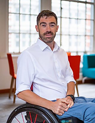 A man smiles at the camera. He is wearing a white shirt and blue trousers. He has short, dark hair and stubble. He is sitting in a black wheelchair, in a large open room with tall windows behind him.