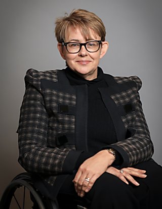 A lady wearing a black outfit including a jacket with prominent shoulder pads, smiles at the camera. She has short brown hair with highlights and black, thick framed glasses. She is sitting in a wheelchair.