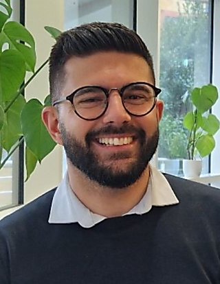A man with well groomed, dark hair smiles at the camera form behind, black, round lense glasses. He has a short beard and is wearing a white shirt, with a navy blue jumper over.