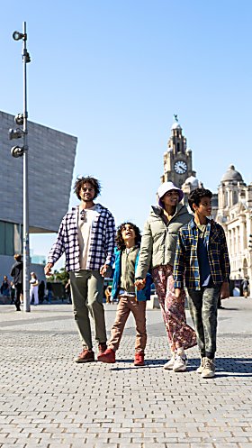 A family of four, two adults and two children walking across Liverpool Pier Head with the Royal Liver Building in the background.