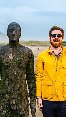 A life-size cast-iron statue on Crosby Beach in a standing position. There is a person standing next to the statue, smiling and wearing a yellow raincoat and sunglasses. They are mimicking the stance of the statue.