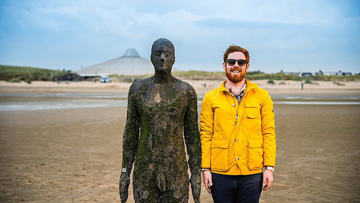 A life-size cast-iron statue on Crosby Beach in a standing position. There is a person standing next to the statue, smiling and wearing a yellow raincoat and sunglasses. They are mimicking the stance of the statue.
