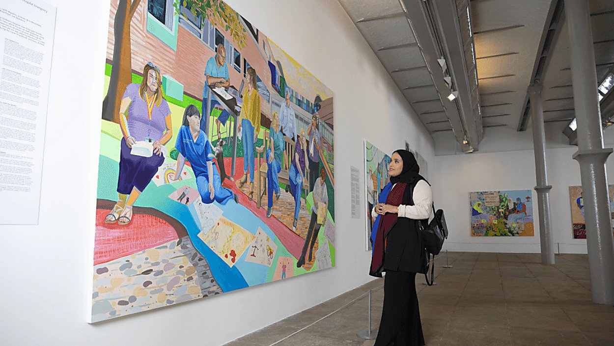 A lady wearing long black clothes looks at a large colourful and lifelike painting in a gallery. The gallery has white walls and grey metal supports.