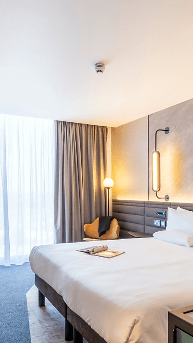 An extra large white bed in a spacious, airy room. The walls are a grey marble on one side and a deep green on the other. To the right is a special coffee machine and nearby a selection of coffee pods.