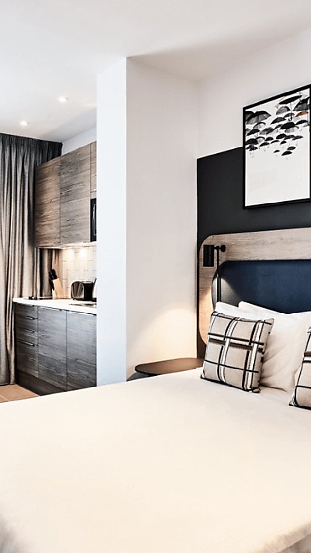 A modern apartment with a large white bed, kitchentte, large window and wall mounted TV.