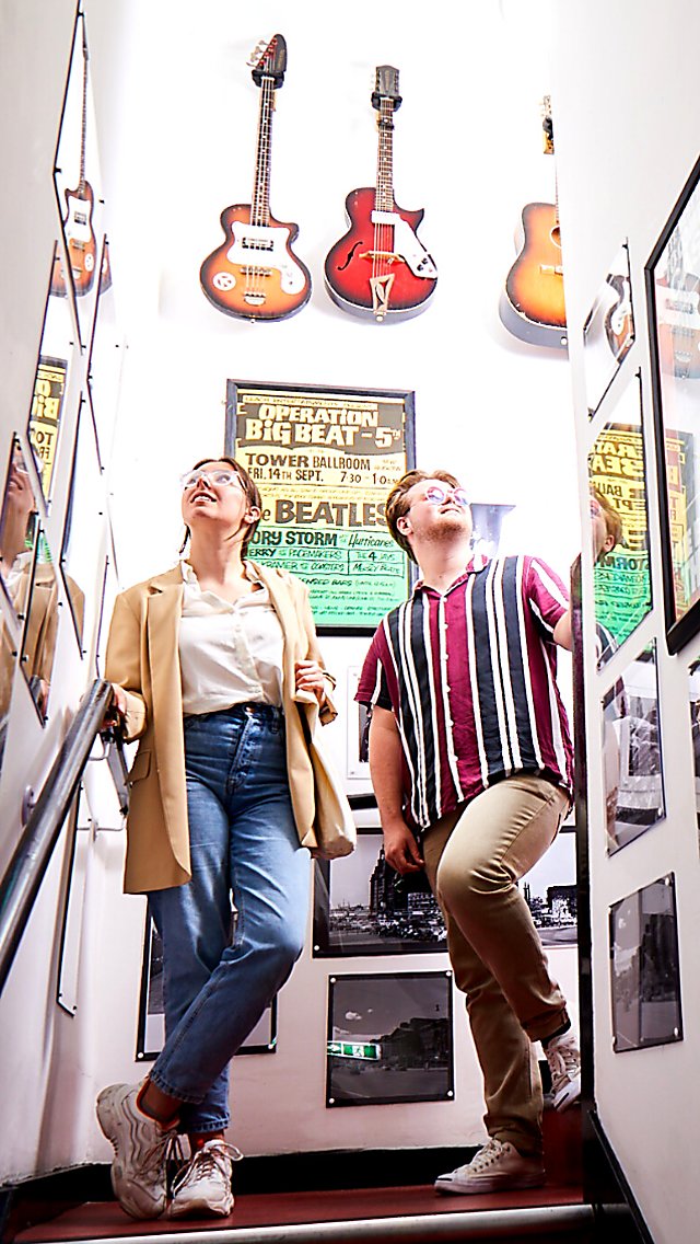Two young people stand in the stairway of the Beatles Museum lined with Beatles posters and guitars.