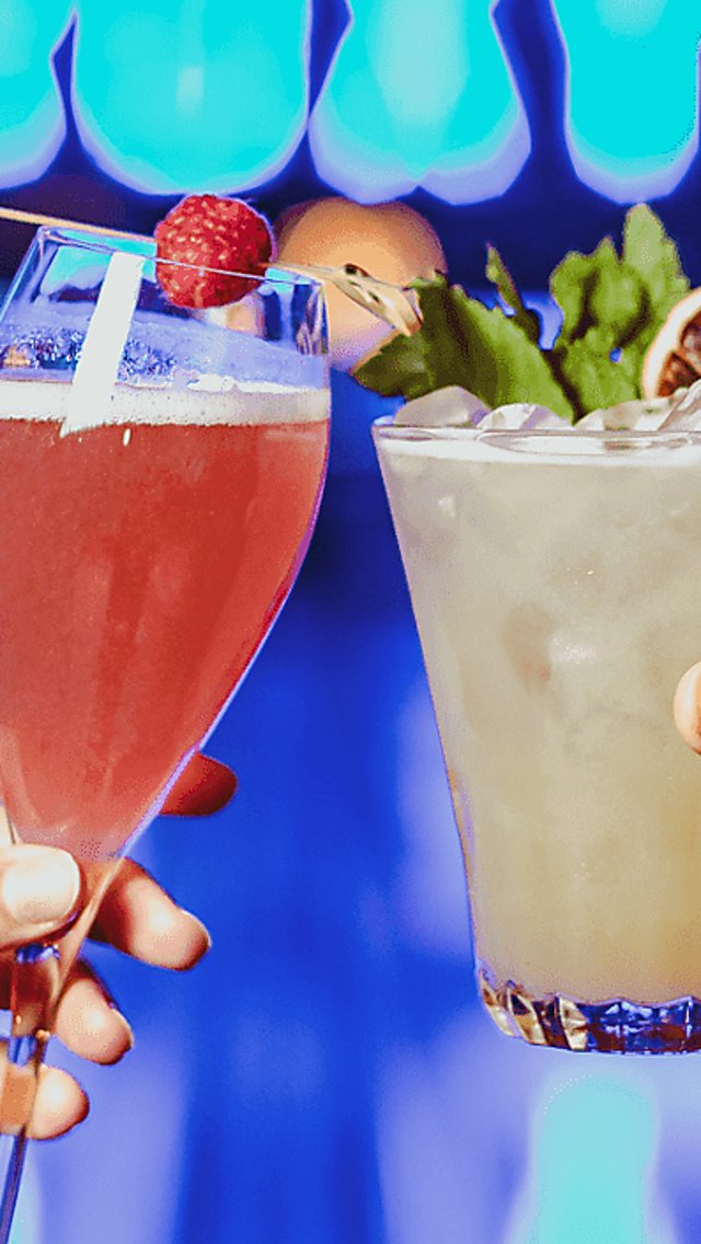 Two people 'cheers'ing two cocktails in front of a neon sign saying 'ROXY'. The left drink is a bellini style cocktail and a red pink colour. The cocktail on the right is a shorter glass with a margarita style cocktail.
