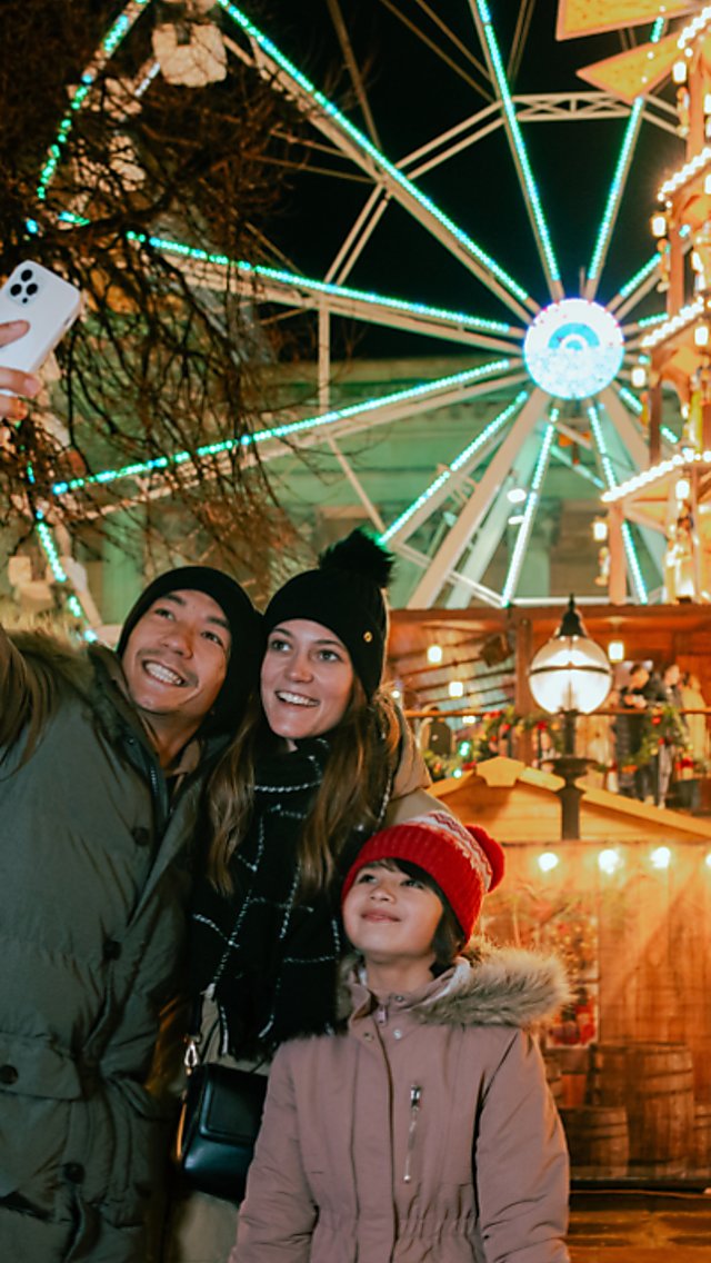 A family of three people, two adults an a child, taking a selfie in front of the Christmas Markets in Liverpool. St George's hall and a large ferris wheel is in the background.