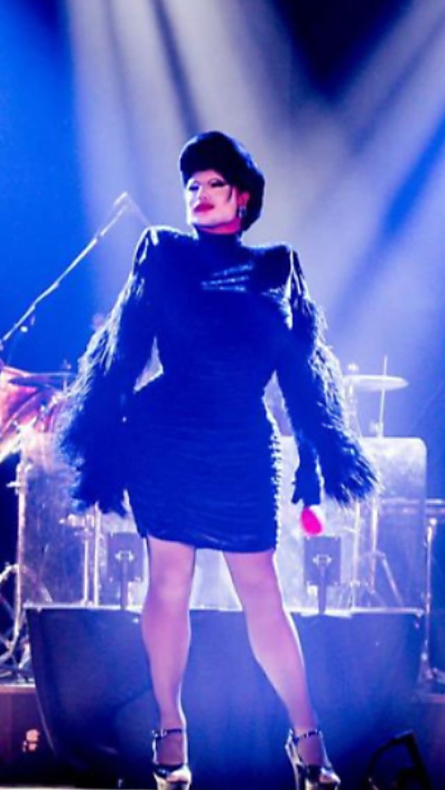Danny Beard on stage in drag surrounded by a band with blue lights on them