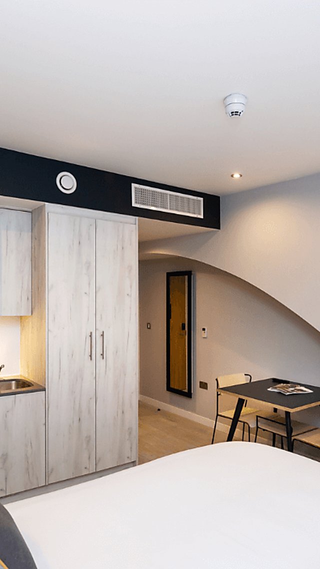 Inside an aparthotel that is airy and bright with a large white bed, a small kitchenette and flat screen TV on a whitewall.