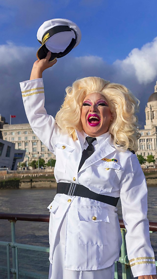 A drag artist on board the Mersey Ferry dressed as a sailor, wearing a white oufit, large blonde wig and saluting their sailor hat. In the background is the Liverpool skyline on a sunny day.