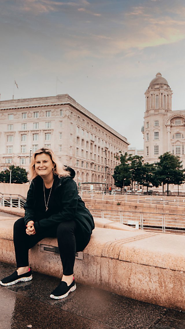 A person dressed in dark clothing sits on a wide stone ledge beside a wet waterfront promenade, with historic Liverpool buildings in the background, including the Royal Liver Building with clock towers on the left and a large domed building on the right. The ground appears rain-soaked, and the scene is lit by soft, overcast daylight with a muted sky.