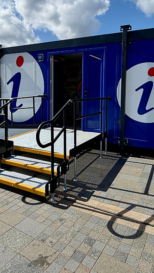 Blue Visitor Welcome Centre cabin on Liverpool’s waterfront with accessible ramp and steps, featuring ‘Visitor Welcome Centre’ and ‘VisitLiverpool.com’ signage, set beside the riverside promenade under a cloudy sky.