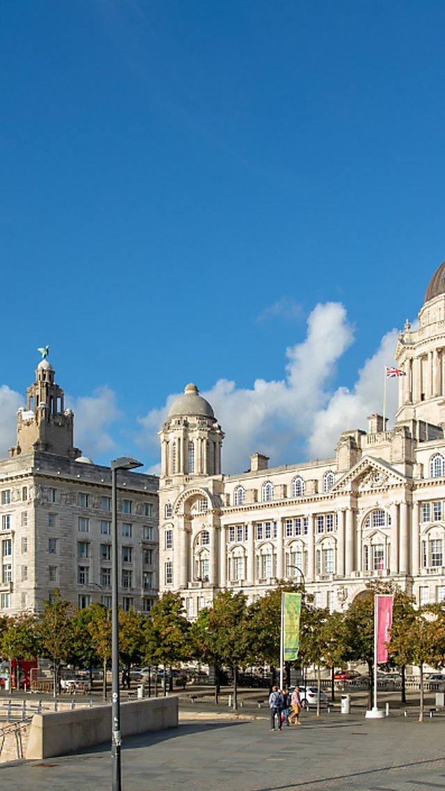 A wide daytime view of Liverpool’s waterfront featuring the Royal Liver Building with its clock tower on the left and the domed Port of Liverpool Building on the right. The historic white stone buildings line a tree-filled plaza with paved walkways, a few pedestrians, and flags on poles. The sky is bright blue with scattered clouds, and modern buildings are visible in the distance.