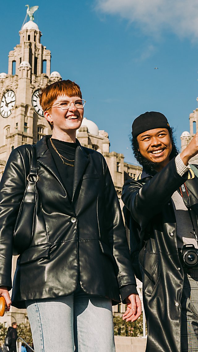 Two people infront of the Royal Liver Building similing. One is pointing at something off camera.