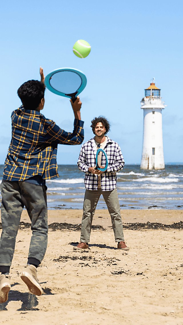 Two people playing tenning on a beach with a lighthouse in the background.
