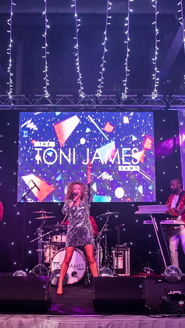 Live band performing on a stage decorated with hanging fairy lights and starry backdrop, with multiple musicians playing instruments and a lead vocalist at centre. A large screen behind the stage displays the text “The Toni James Band,” with purple and blue stage lighting and Christmas trees positioned at either side.