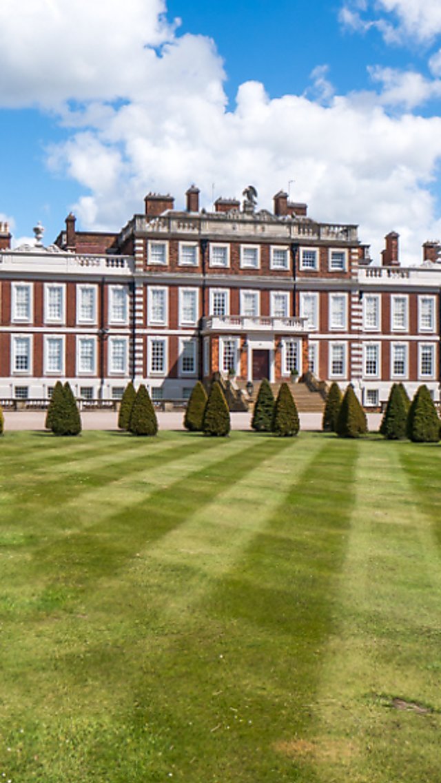 Wide view of a grand historic country house with red-brick and stone façades, set behind a neatly striped lawn and a row of conical trimmed trees, with purple wisteria in bloom along the left side and a bright blue sky with scattered clouds overhead.