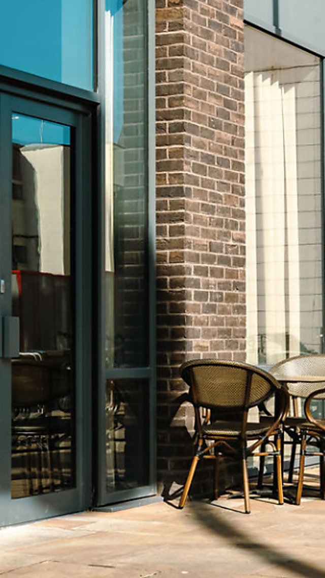 Exterior of a contemporary brick building with large glass doors and windows, a sign reading “Clubhouse” beside the entrance, and outdoor café-style tables and chairs arranged on a paved terrace.
