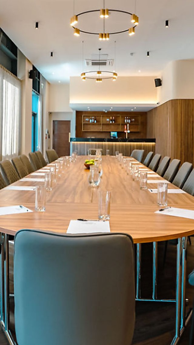Modern boardroom with a long oval wooden table set with notepads, pens, water glasses and jugs, surrounded by upholstered chairs, with floor-to-ceiling curtains and warm pendant lighting overhead.