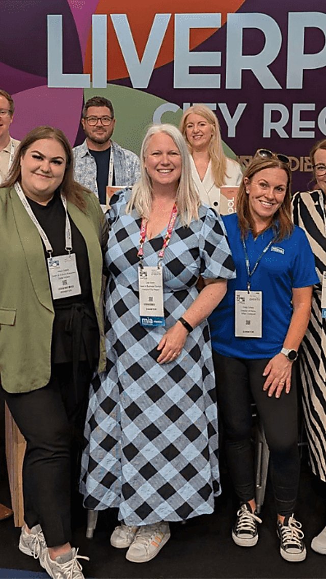 A group of people standing looking at the camera and smiling brightly. There is a sign behind them which says ''LIVERPOOL CITY REGION''.
