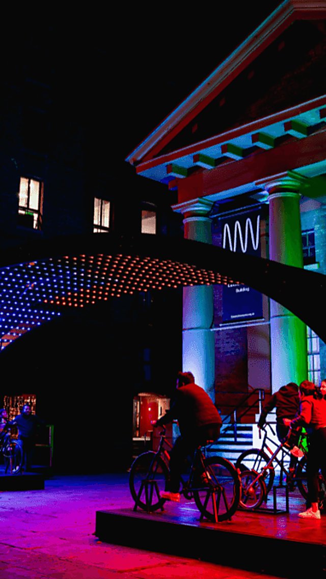 Purple and green lights illuminating the area with people on bikes as part of the installation.