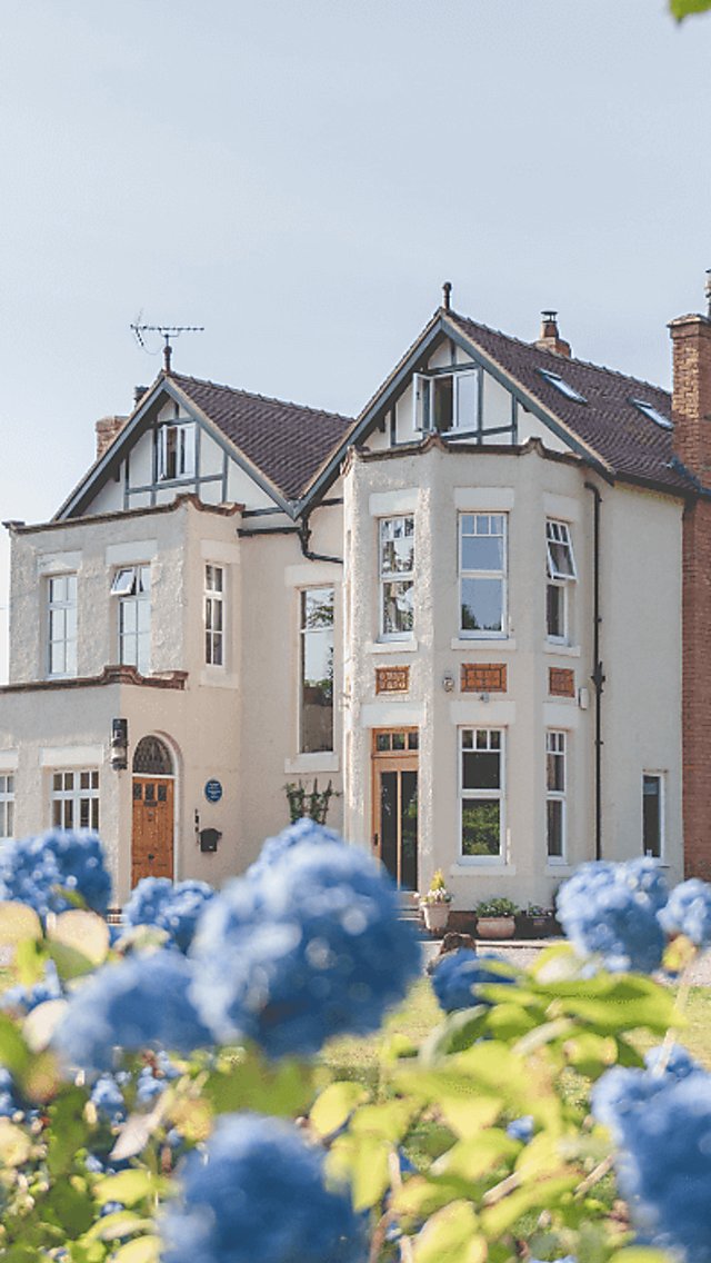 A large manor house painted white with huge windows and two pointed rooves. The photo is taken amongst blue flowers on a sunny day.