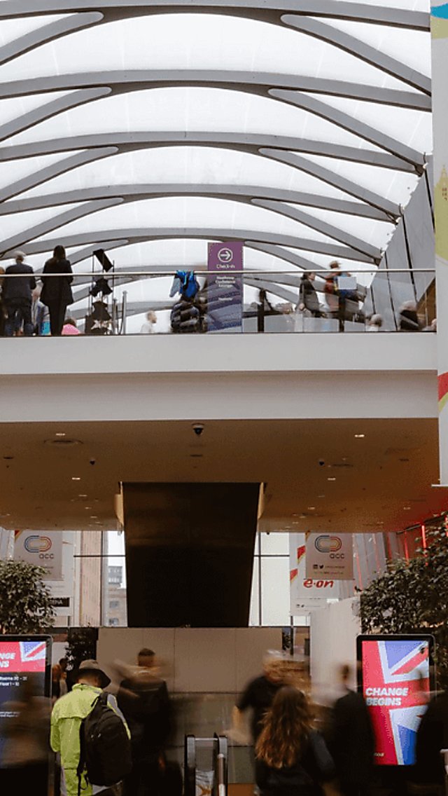 A conference taking place in ACC Liverpool, there are people walking around talking to one another with banners on either side of the wall, hanging from above.