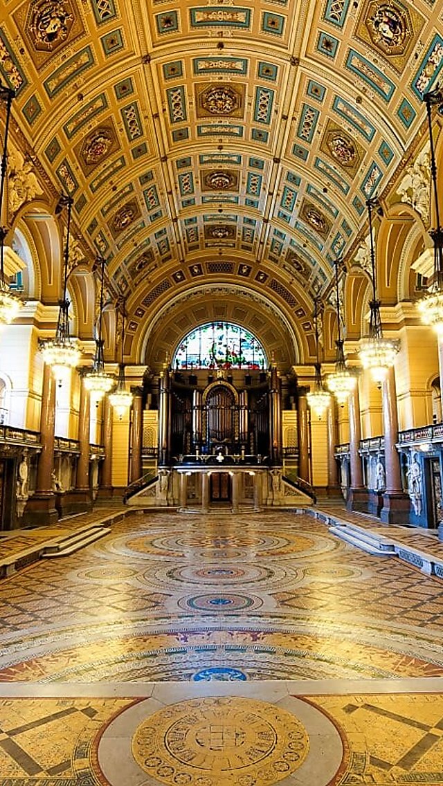 The Main Hall at St George's Hall with an ornate vaulted ceiling, decorative chandeliers, and tall marble columns lining both sides. The space features an intricate patterned Minton tile floor leading toward a large pipe organ and stained-glass window at the far end. Warm lighting highlights the detailed architecture, carved statues, and elaborate decorative finishes throughout the hall.