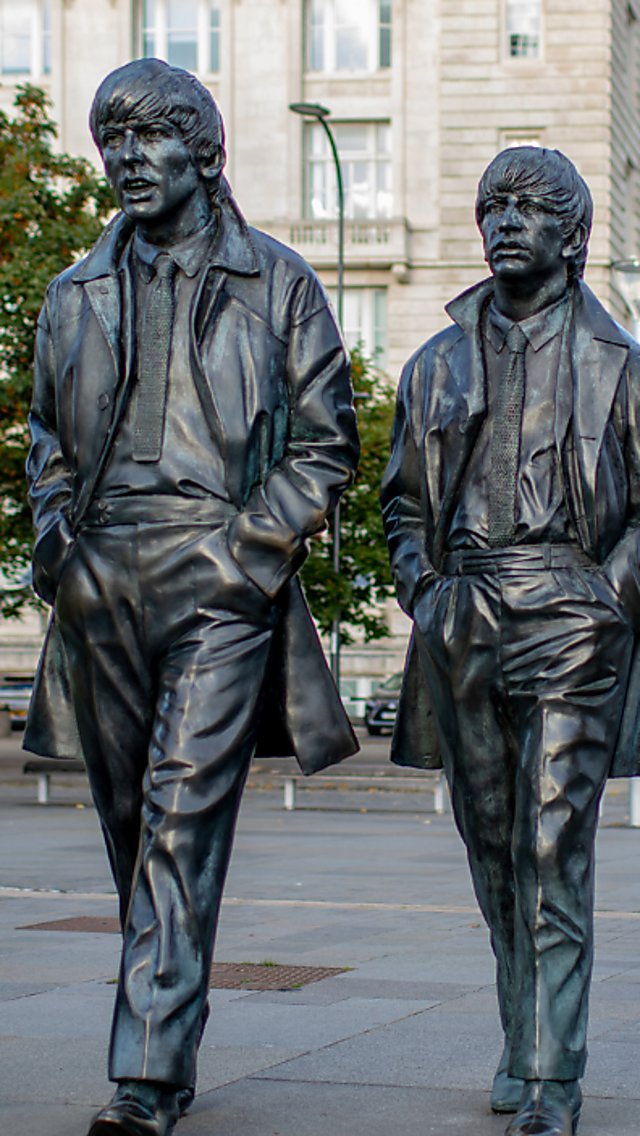 Bronze statue of four walking figures in a city plaza, each wearing coats and mid‑20th‑century clothing. One figure holds a coat while the others have hands in pockets. The sculptures stride forward side by side on a paved walkway, with a large stone building, trees, and modern city structures in the background under an overcast sky.
