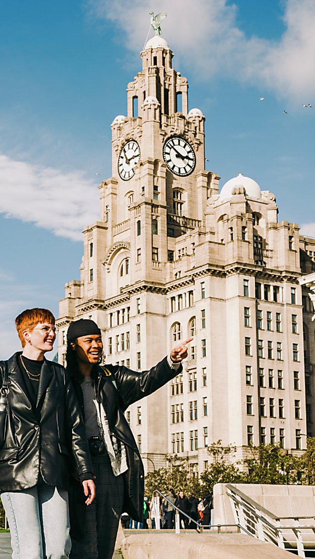 Two people walking past the royal liver building in Liverpool on a sunny day. One of the people is pointing to something to the right of camera.