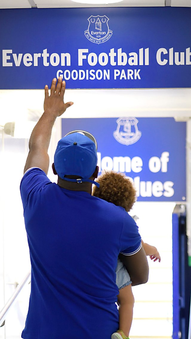 A person carrying a small child walking down the players tunnel at Goodison Park. they are touching the Everton sign above their head.