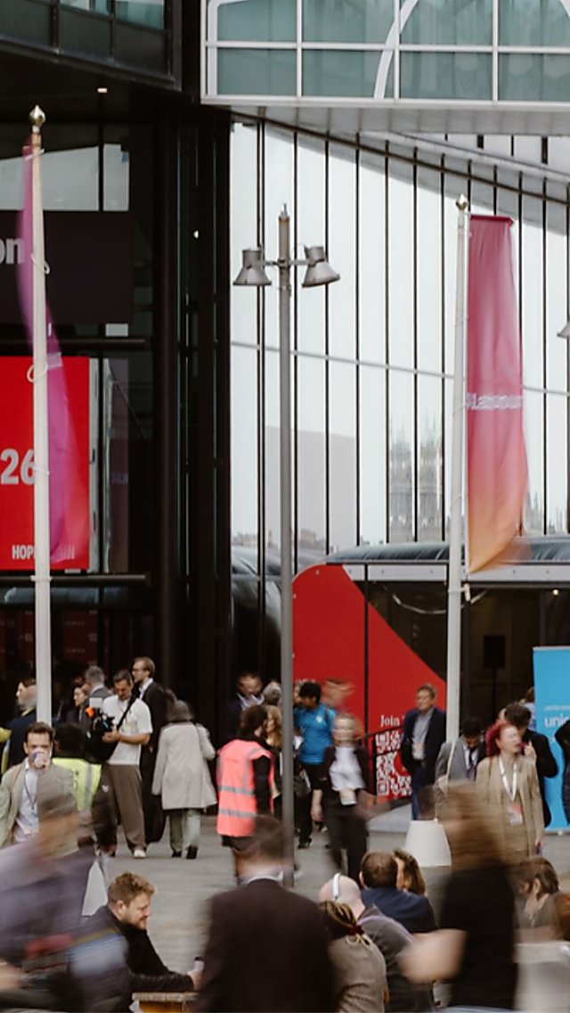 Busy crowds arrive and gather outside a modern exhibition centre, where a large red digital screen displays “Welcome to Conference 2026.” Glass-fronted buildings, banners and overhead walkways frame the scene, highlighting the scale and activity of the event.
