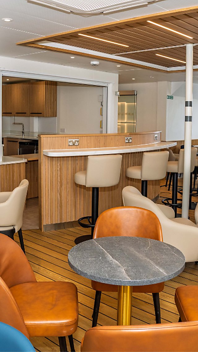 Modern indoor lounge on a ferry or ship with light wood flooring and wood-accented walls, featuring a central bar counter with high stools. Around the room are small round tables surrounded by cushioned armchairs in beige, blue, and tan. Large rectangular windows on the right provide a view of a city skyline over water. Overhead, slatted wooden ceiling panels and recessed lighting create a warm, contemporary atmosphere. Visible signage on the left indicates a muster station.