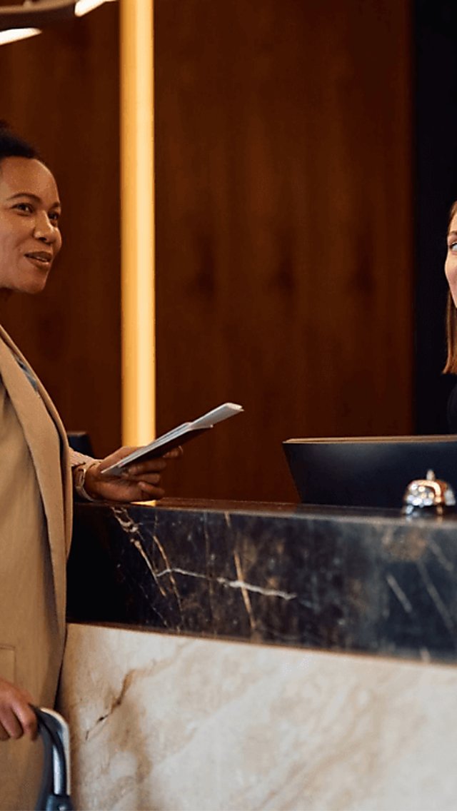 Two people standing at a reception desk with a receptionist standing behind the desk advising the person.