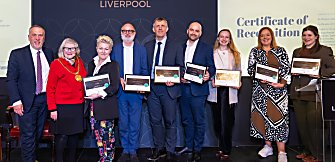 Group of people standing on a stage holding certificates, posed in front of a ‘Club Liverpool’ backdrop with a ‘Certificate of Recognition’ screen displayed behind them.