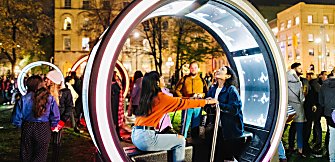 Two people sitting in a round light artwork looking at the roof of it. They are holding onto a bar in the middle of the artwork.