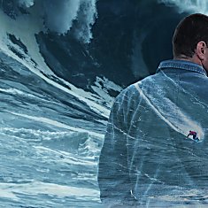 A creative double-exposure style image showing a person in a blue jacket viewed from behind, set against a dramatic ocean scene with towering waves. A small surfer is depicted riding a wave within the silhouette of the jacket, blending the person and seascape together. The image uses cool blue tones and textured water effects to create a striking, artistic composition.