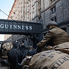 A large brick building with a sign that says Guinness on it and horse and carts pulling large sacks.