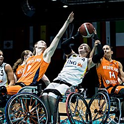 Several athletes in sports wheelchairs compete for a basketball on an indoor court. Two athletes in bright orange jerseys with “NEDERLAND” visible and one in a white jersey with “ING” reach upward toward the ball at the same moment, arms extended. Additional players in white and orange uniforms cluster closely around them. The scene is tightly framed on the action, with visible wheelchair frames, angled wheels, and protective gear. In the background, a row of international flags hangs above the court under dim arena lighting.
