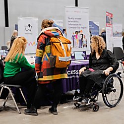 One able bodied person standing with another sitting on a chair and taking to a wheelchair user at the market place.