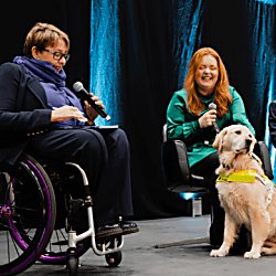 A guide dog user sitting on stage with a wheelchair user who is looking at an iPad.
