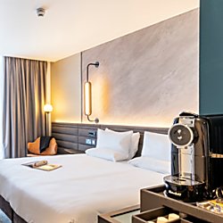 An extra large white bed in a spacious, airy room. The walls are a grey marble on one side and a deep green on the other. To the right is a special coffee machine and nearby a selection of coffee pods.